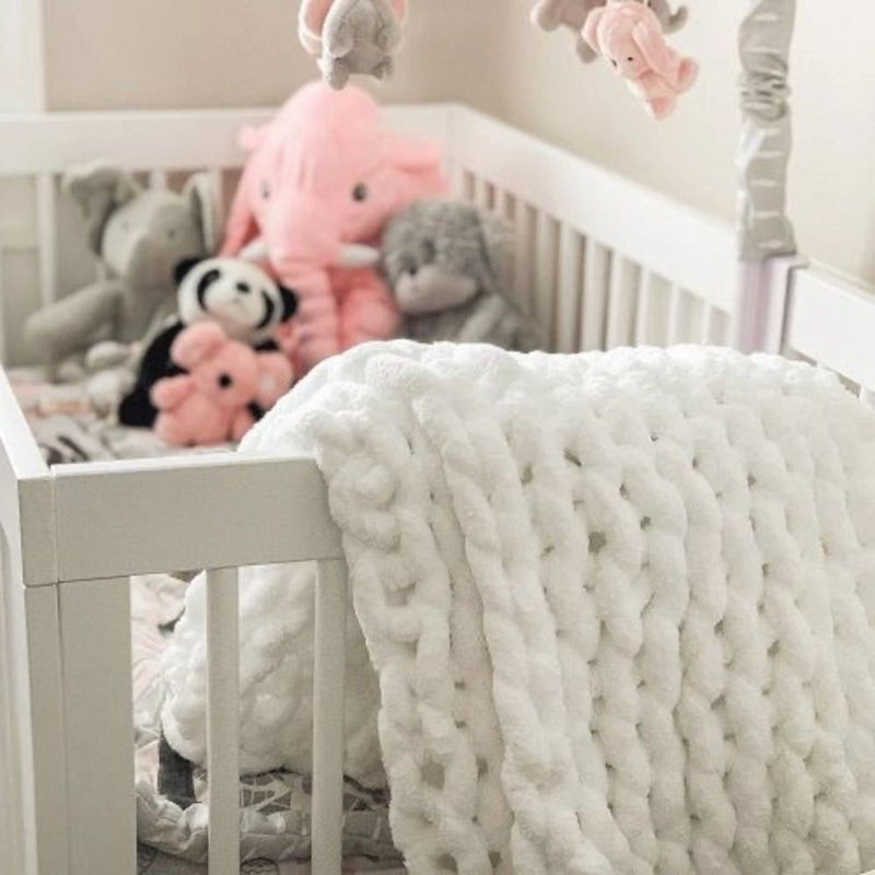 White baby cot with grey & pink plush toys featuring a white chunky knitted baby blanket